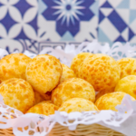 Cesta com pães de queijo dourados e recém-assados sobre guardanapo rendado, com fundo de azulejos azuis decorativos.