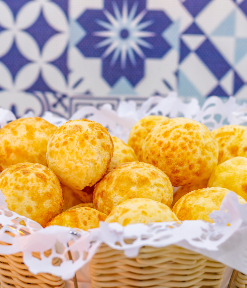 Cesta com pães de queijo dourados e recém-assados sobre guardanapo rendado, com fundo de azulejos azuis decorativos.